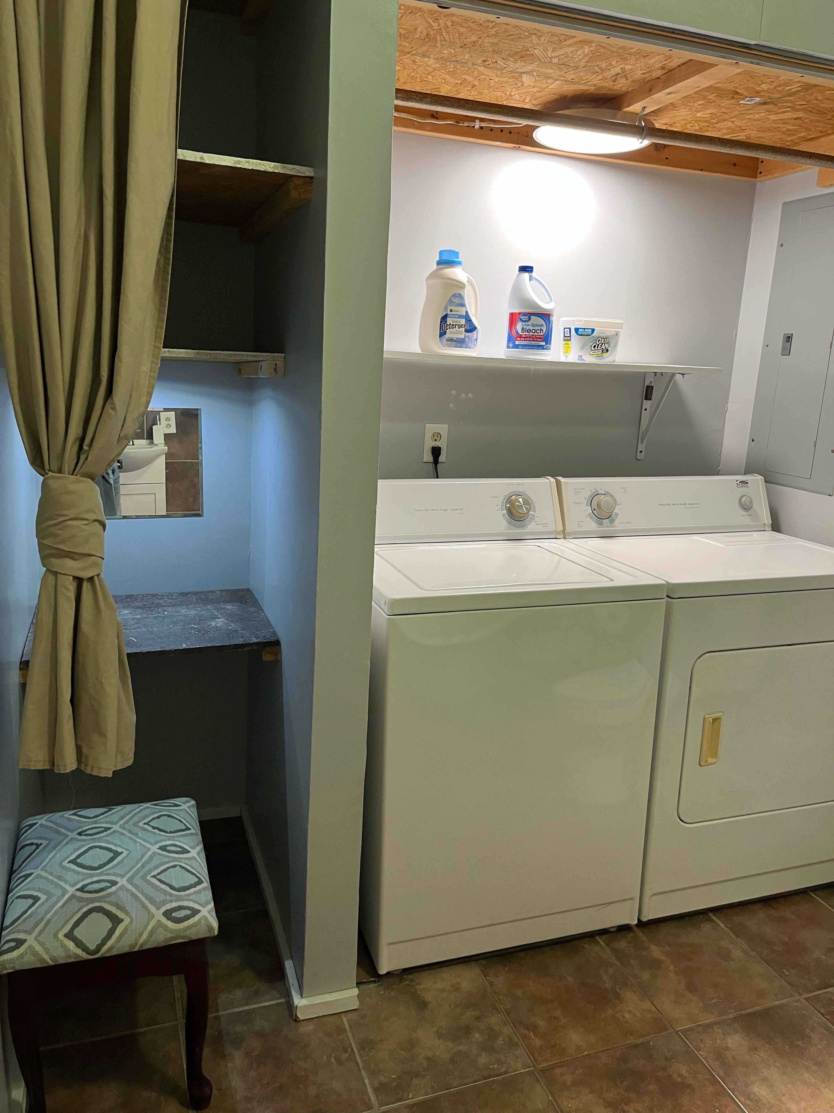 A laundry room with a washing machine, detergent bottles, and a small bench.
