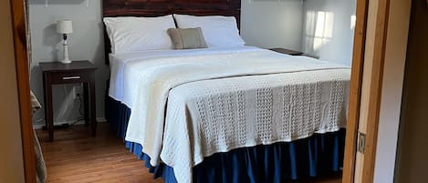 Un dormitorio con piso de madera, una cama con cubrecama blanco y azul, un ventilador de techo y estantes empotrados.