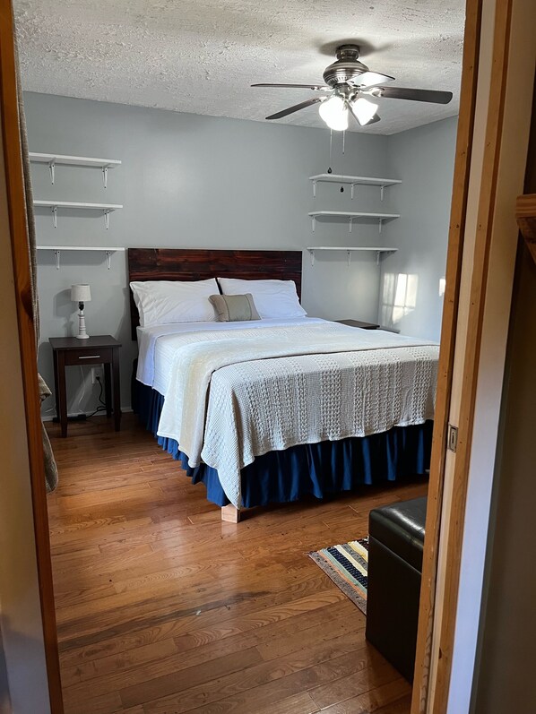 Une chambre avec un plancher en bois, un lit recouvert d’une courtepointe blanche et bleue, un ventilateur de plafond et des étagères intégrées.