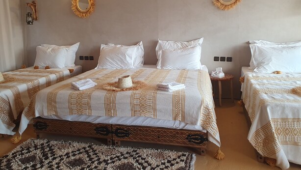 A hotel room with two beds, a large bed with a patterned bedspread and a smaller bed with a plain bedspread. There are two wall-mounted lamps and two wall-mounted mirrors.