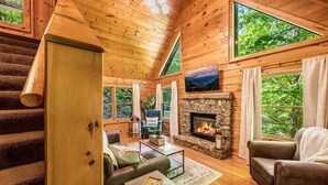 A cozy cabin interior with a stone fireplace, wooden floors, a sofa, a coffee table, and large windows offering views of the surrounding forest.