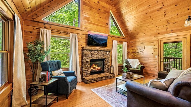 A cozy cabin interior with a stone fireplace, wooden walls, and large windows offering a view of the greenery outside.