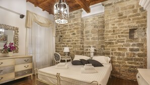 A bedroom with a stone wall, a bed with white linens, a bedside table with a lamp, and a dresser with drawers.
