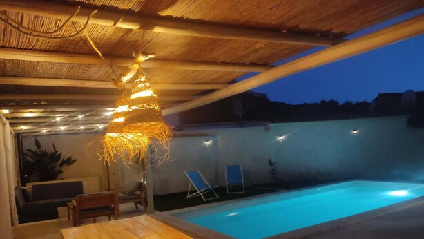 An outdoor pool area with a wooden table and chairs, a hanging light fixture, and two lounge chairs. - Villa les volets bleus - swimming pool - near the calanques of Cassis (Roquefort-la-Bédoule)