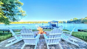 Une terrasse au bord du lac, avec des chaises Adirondack disposées face à un foyer extérieur, un quai et une zone abritée en arrière-plan.