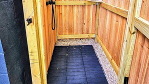 Une douche extérieure en bois, dotée d’un sol en caoutchouc noir, entourée de murs en bois et reposant sur une base de gravier.