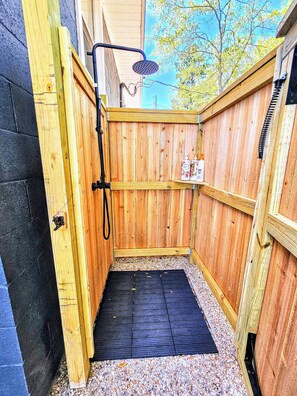 Una ducha exterior de madera con suelo de goma negra, rodeada de paredes de madera y una base de grava.