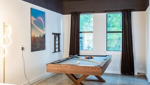Una mesa de billar en una habitación con ventanas amplias, una escalera y un cuadro en la pared.