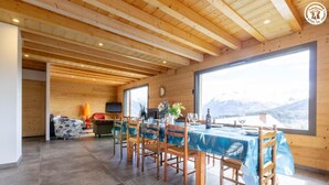 Un comedor con una mesa dispuesta para cuatro, sillas de madera, un televisor y vistas a montañas a través de amplios ventanales.