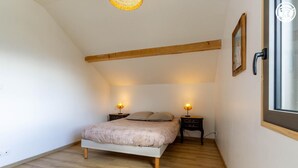Un dormitorio con techo inclinado, una cama con manta color beige, dos mesitas de noche con lámparas y una ventana con marco de madera.
