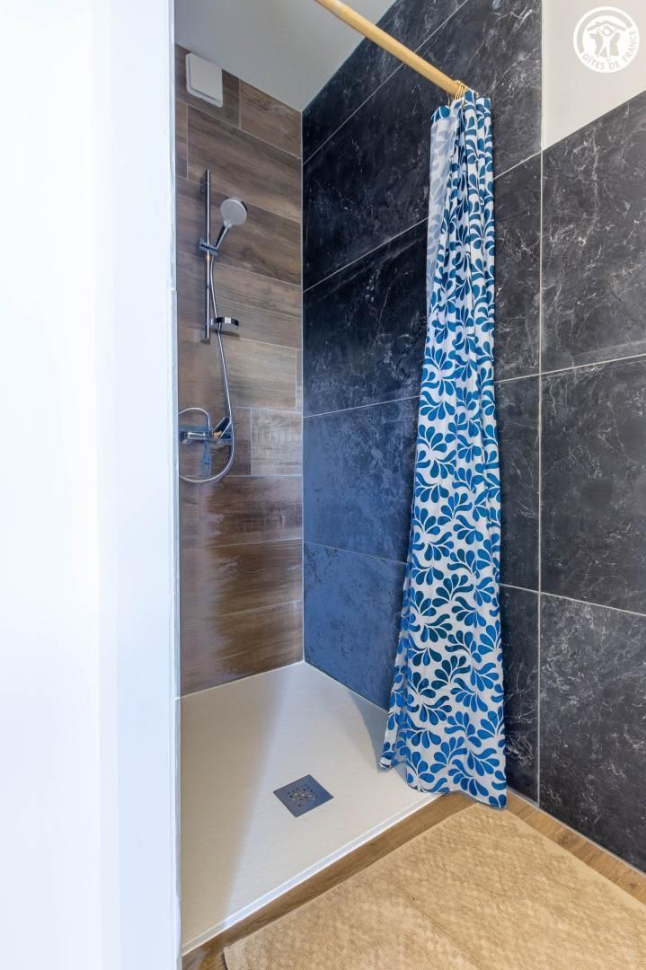 A shower area with a wooden floor, a black tiled wall, and a blue patterned shower curtain.