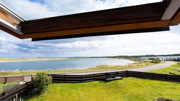 A lakeside view from a modern building with a wooden deck overlooking a calm lake and distant buildings.