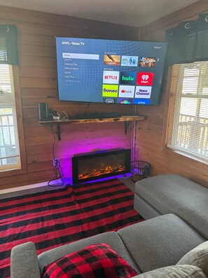 Una sala de estar con una televisión instalada mostrando la interfaz de Roku TV, una chimenea encendida y un sofá gris.