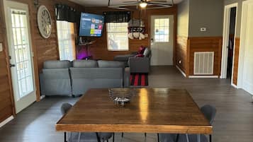 Un salón espacioso con una mesa de comedor de madera, sofás grises y un televisor montado en la pared.