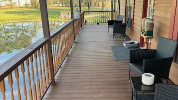 Una terraza de madera con barandilla, mobiliario exterior y vistas a un cuerpo de agua y árboles.