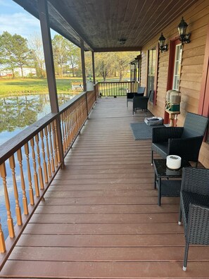 Una terraza de madera con barandilla, mobiliario exterior y vistas a un cuerpo de agua y árboles.