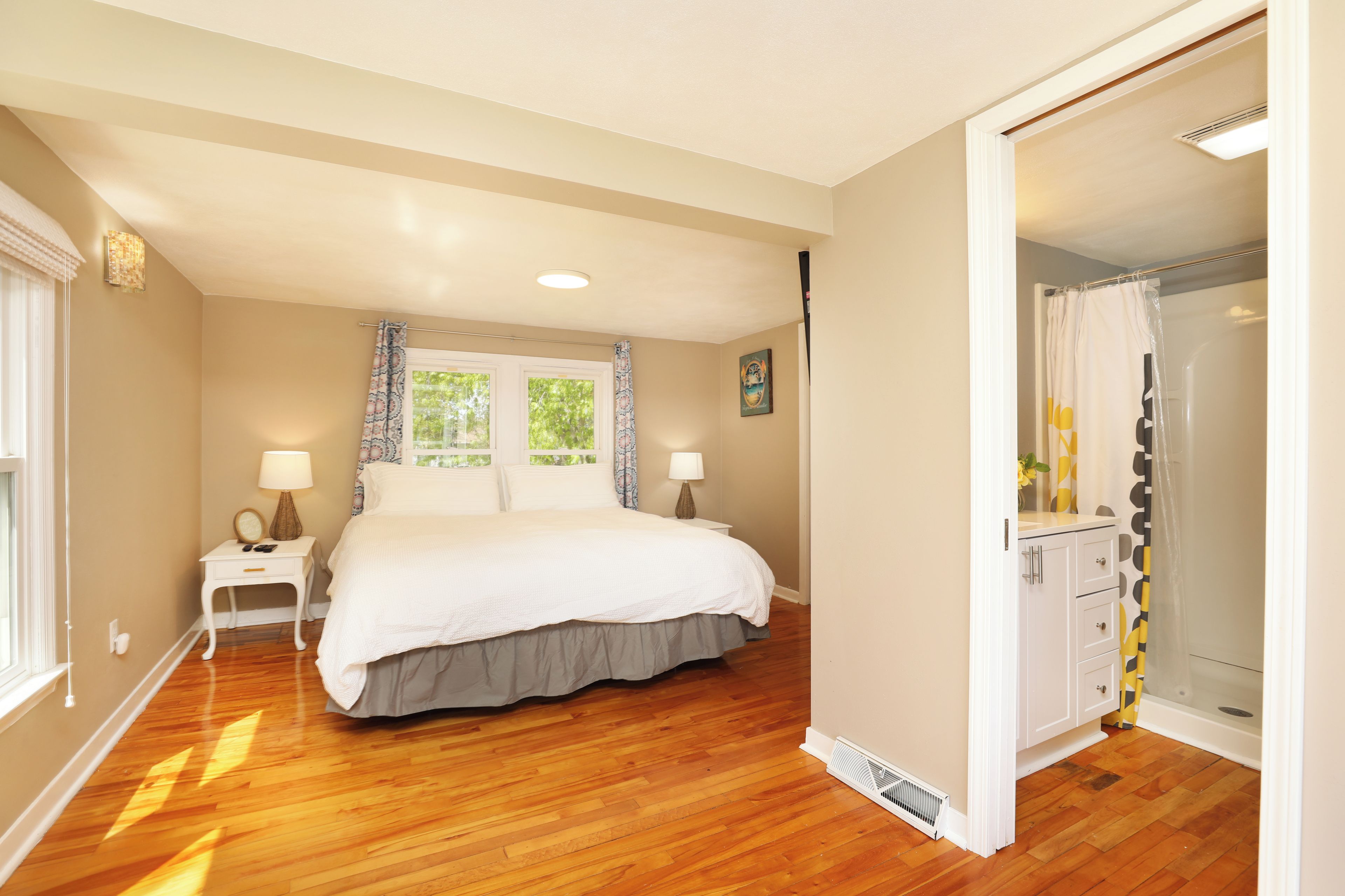 A bedroom with a large bed, a nightstand, a lamp, a window with curtains, and a bathroom with a shower and toilet.