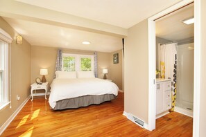 Un dormitorio con una cama grande, una mesita de noche, una lámpara, un ventanal con cortinas y un baño con ducha e inodoro.