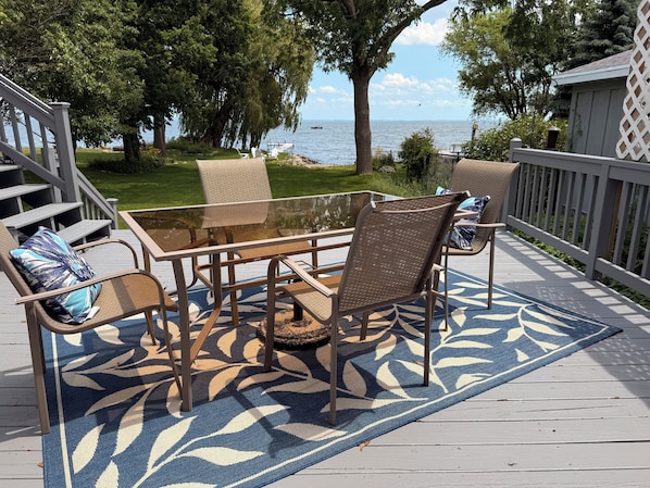 A deck with a glass-top table and wicker chairs, a patterned rug, and a view of the sea.