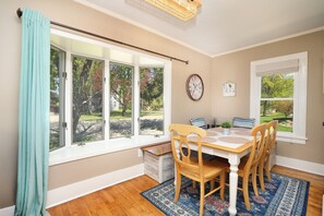 Un comedor con una mesa preparada para cuatro, un reloj en la pared y vistas a un jardín a través de amplios ventanales.