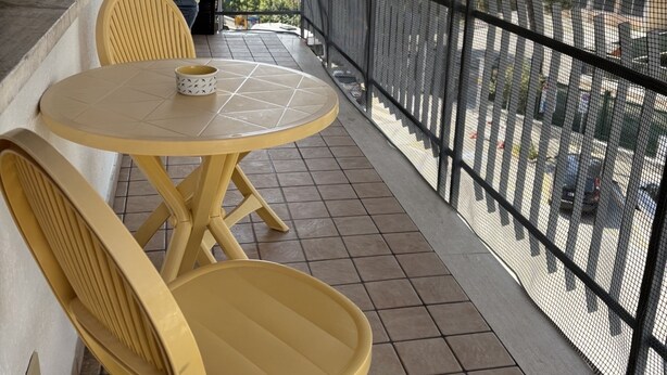 A balcony with a round table and two yellow chairs, a white ceiling fan, and a view of the street below.