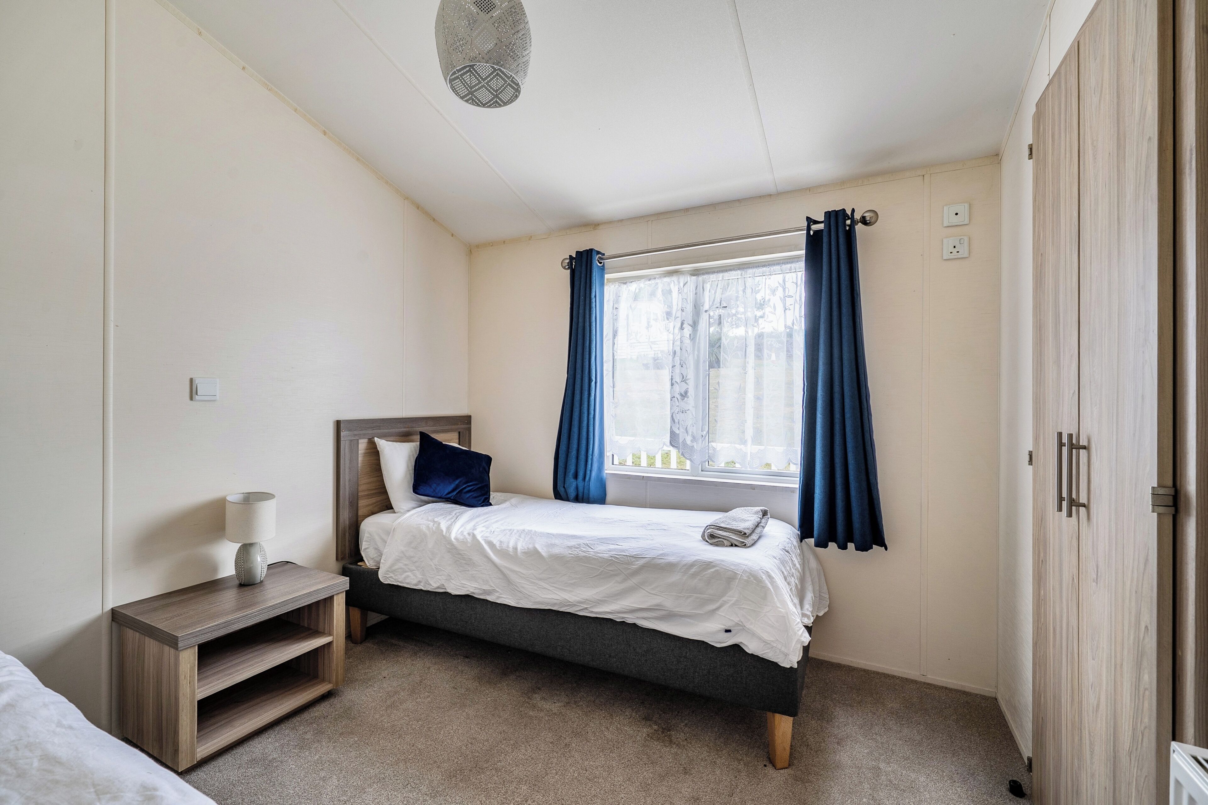 A bedroom with a bed, a nightstand, a lamp, a window with curtains, and a wardrobe.