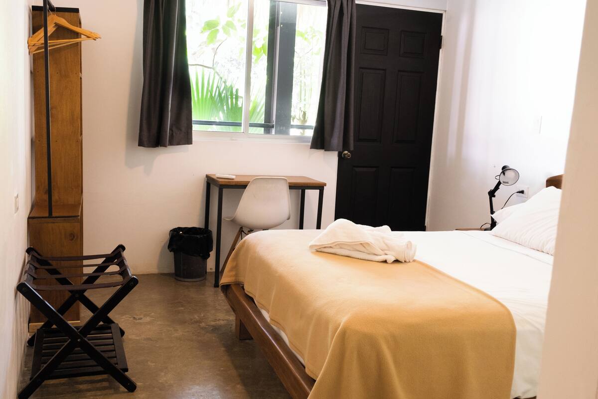 A bedroom with a bed, a desk, a chair, a wooden bench, and a window with curtains.