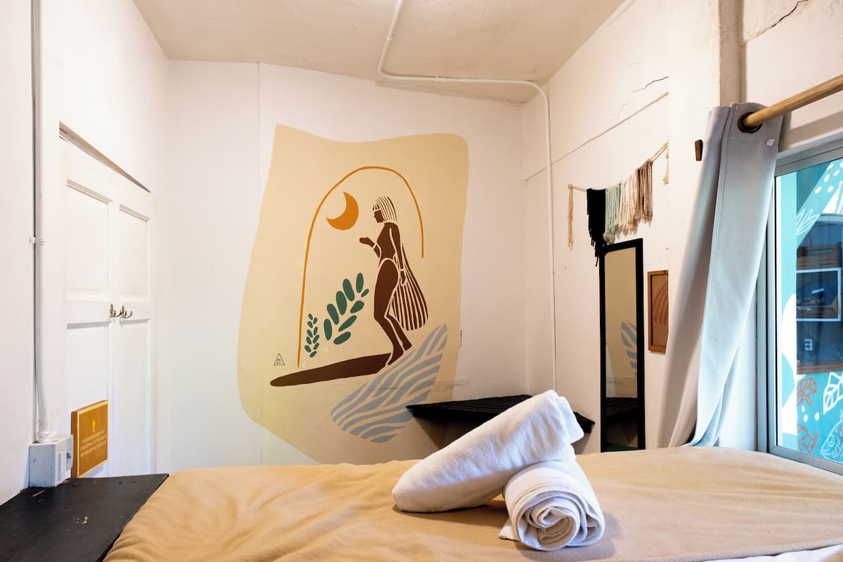 A room with a bed, a towel, a wall mural, a window, and a door.