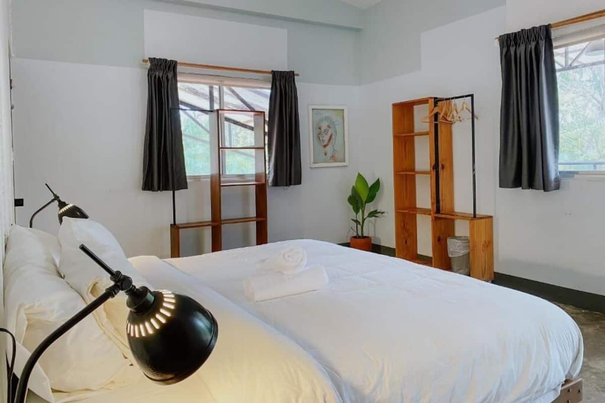 A bedroom with a bed, a bedside lamp, a window with curtains, a wooden shelf, and a potted plant.