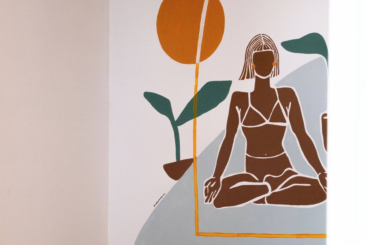 A bed with white pillows and a blanket, a wall with a mural of a woman meditating, and a partial view of a window.