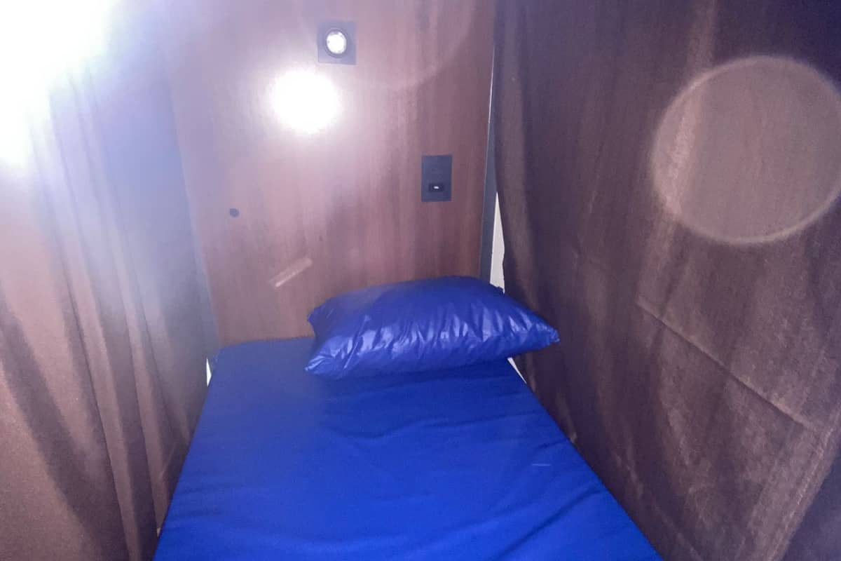 Una stanza piccola e stretta con un letto singolo, un comodino e una lampada fissata al muro.