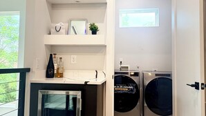 A compact laundry room with a built-in washer and dryer, a small refrigerator, and shelves with decorative items. - Trinity Two Luxury Home (Dallas)