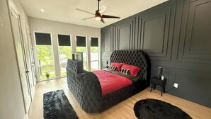 A bedroom with a large bed, a ceiling fan, and a view of greenery outside. - Trinity Two Luxury Home (Dallas)