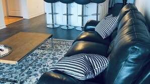 Un salon moderne avec un canapé en cuir noir, un tapis à motifs et un coin cuisine doté de meubles blancs et d’un four à micro-ondes.