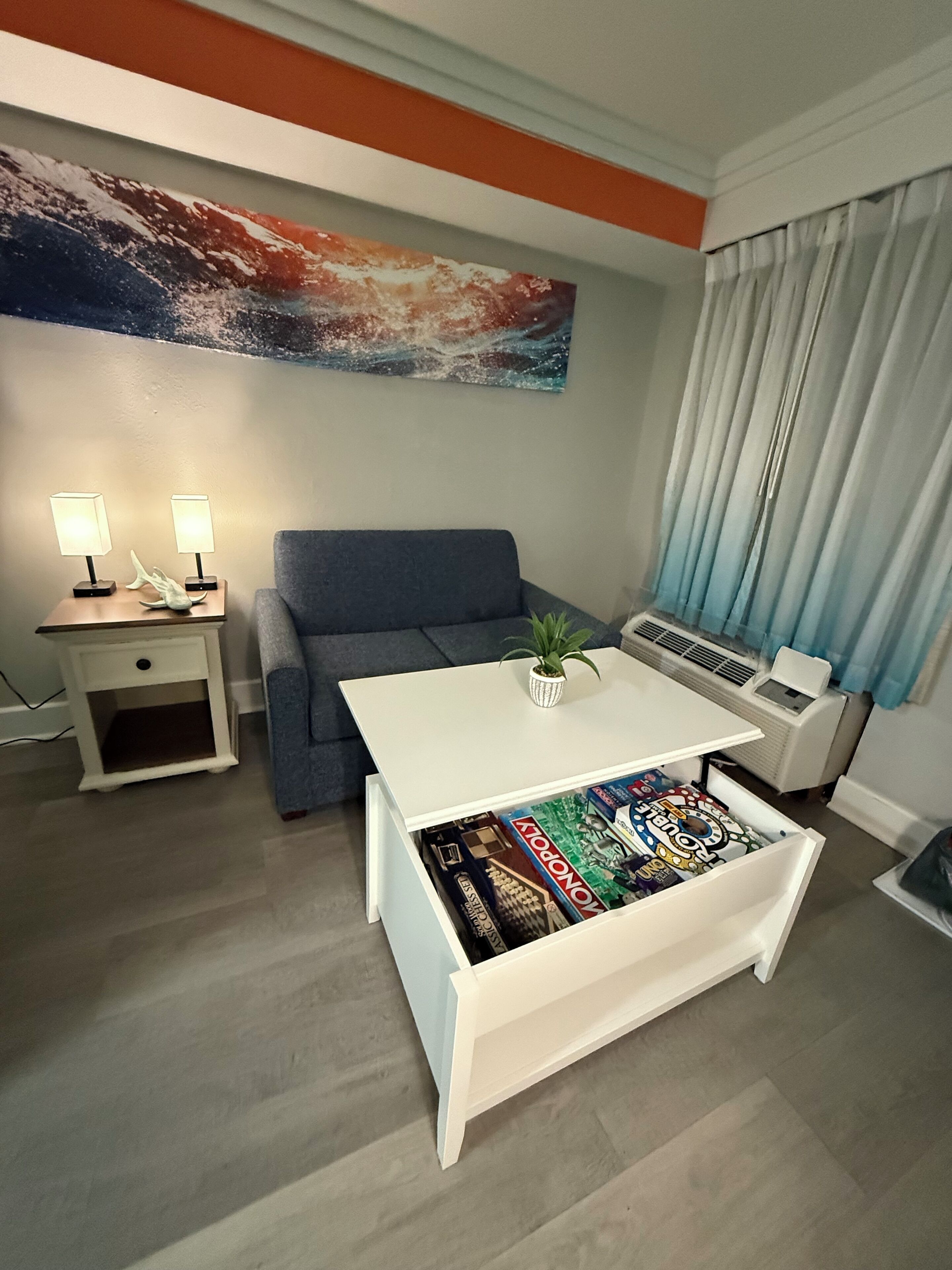 Una sala de estar con un sofá gris, una mesa de centro blanca con juegos de mesa, una mesita de noche con lámparas y un cuadro abstracto grande en la pared.