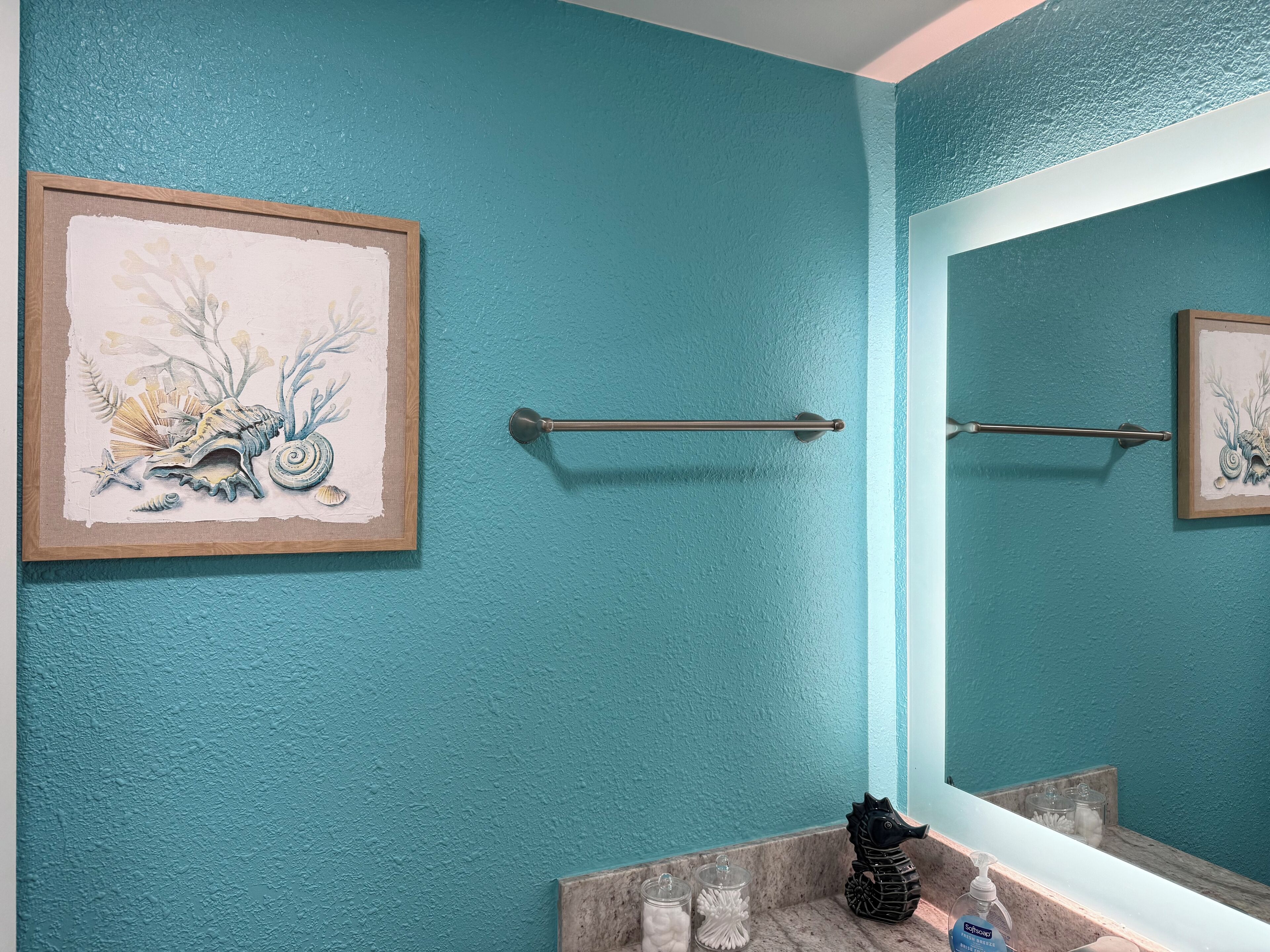 Une salle de bain aux murs de couleur turquoise, ornée d’un tableau encadré représentant une scène de bord de mer, d’un porte-serviettes, d’un miroir et d’une décoration en forme d’hippocampe.
