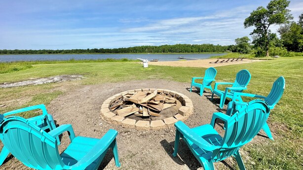 A fire pit with chairs around it, a sandy beach, and a lake in the background.