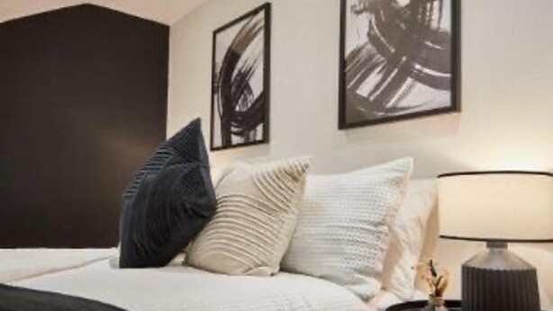 Una cama bien hecha con edredón blanco y gris, una almohada gris oscuro y una almohada blanca. Hay una lámpara negra sobre una mesita de noche.