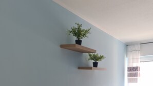 Una habitación con una mesa y silla de madera, dos estantes en la pared con plantas en macetas, y vistas al exterior a través de una ventana con cortinas.
