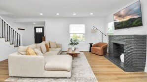 A modern living room with a beige sectional sofa, a stone fireplace, a flat-screen TV, and a staircase leading to another room. - Media Room & Patio - Downtown View- 4 Bedrooms (Houston)