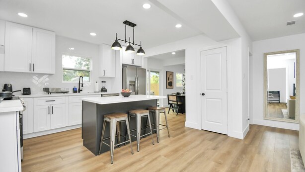 A modern kitchen with white cabinetry, a central island with stools, and a dining area visible through an open door. - Media Room & Patio - Downtown View- 4 Bedrooms (Houston)