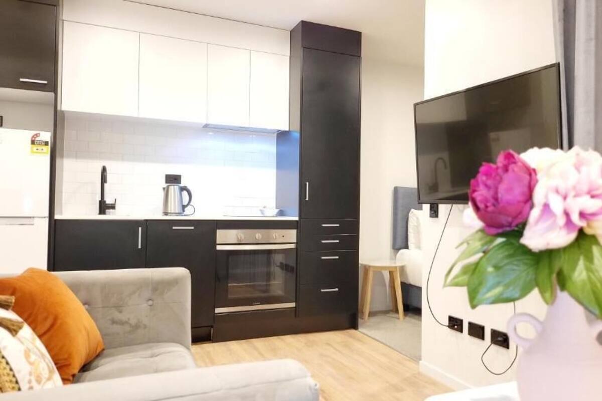 Un moderno salón con un sofá gris, una mesa de centro y un jarrón con flores. Se ve una cocina con armarios oscuros y un televisor al fondo.