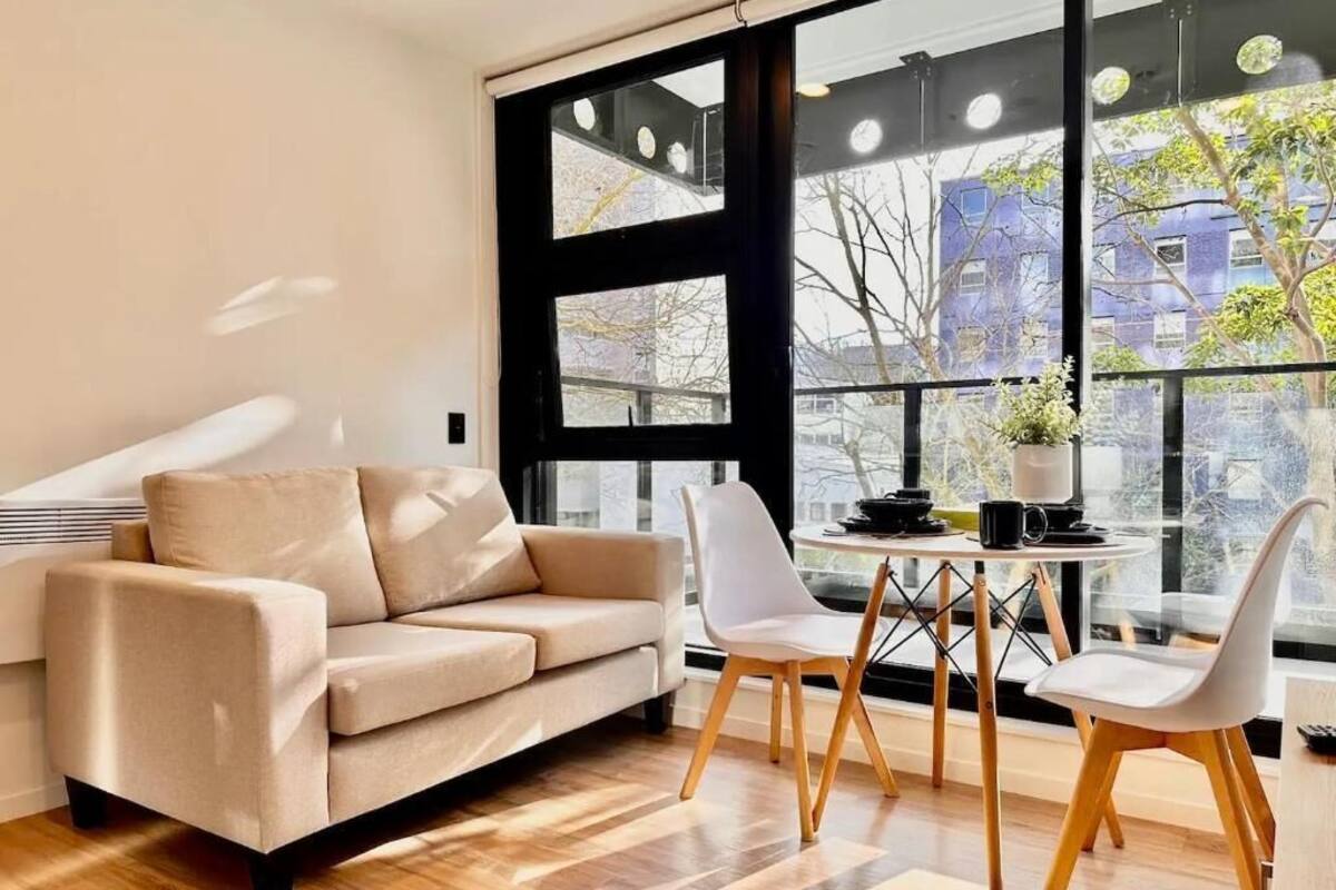 Un salón moderno con un sofá color beige, dos sillas blancas, una mesita redonda y grandes ventanales con vistas a edificios y árboles.