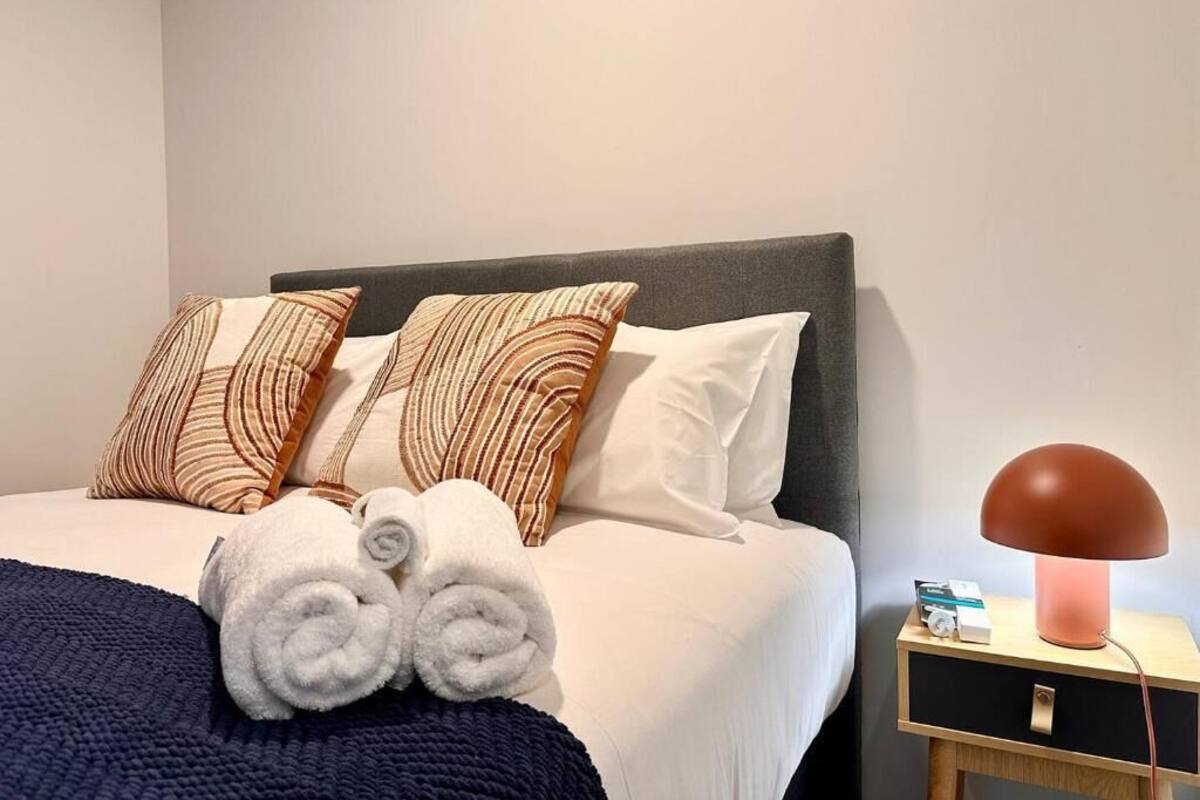 Una cama bien hecha con almohadones estampados, un par de toallas enrolladas y una lámpara de noche sobre una mesita de madera.