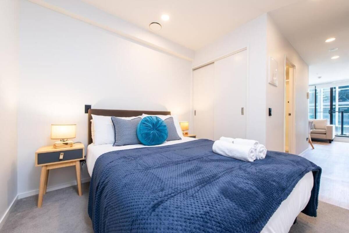 Una habitación de hotel moderna con una cama grande, una mesita de noche con una lámpara y vistas a la ciudad a través de la ventana.
