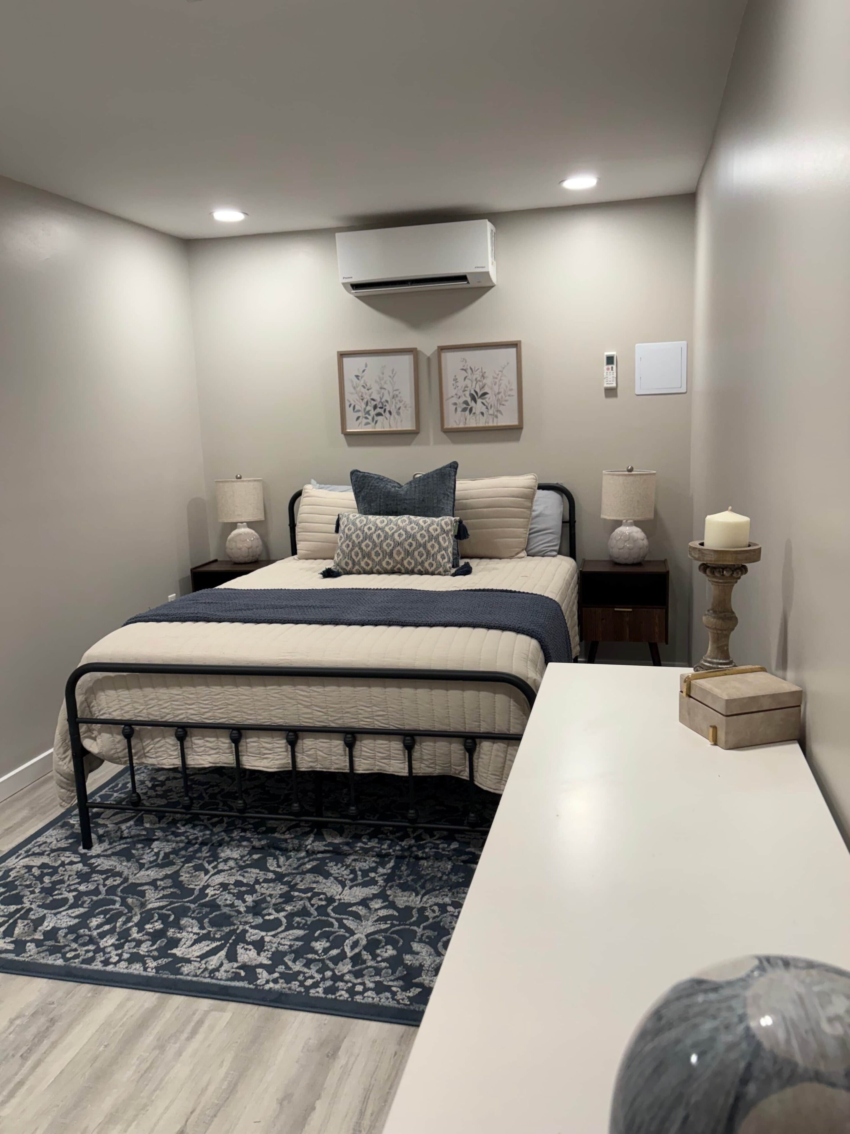 Un dormitorio con cama, mesitas de noche, escritorio, lámpara, vela y aire acondicionado.