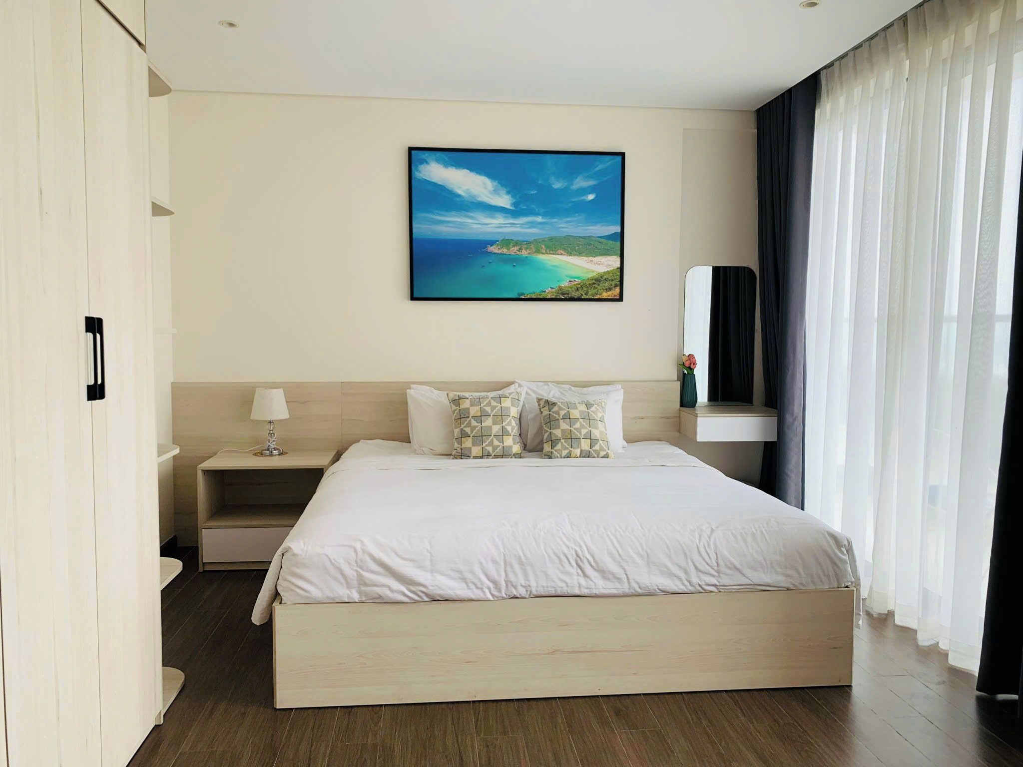 Ein modernes Hotelzimmer mit einem großen Bett, einem Nachttisch, einem Gemälde einer Küstenlandschaft, einem Spiegel und durchsichtigen Vorhängen.