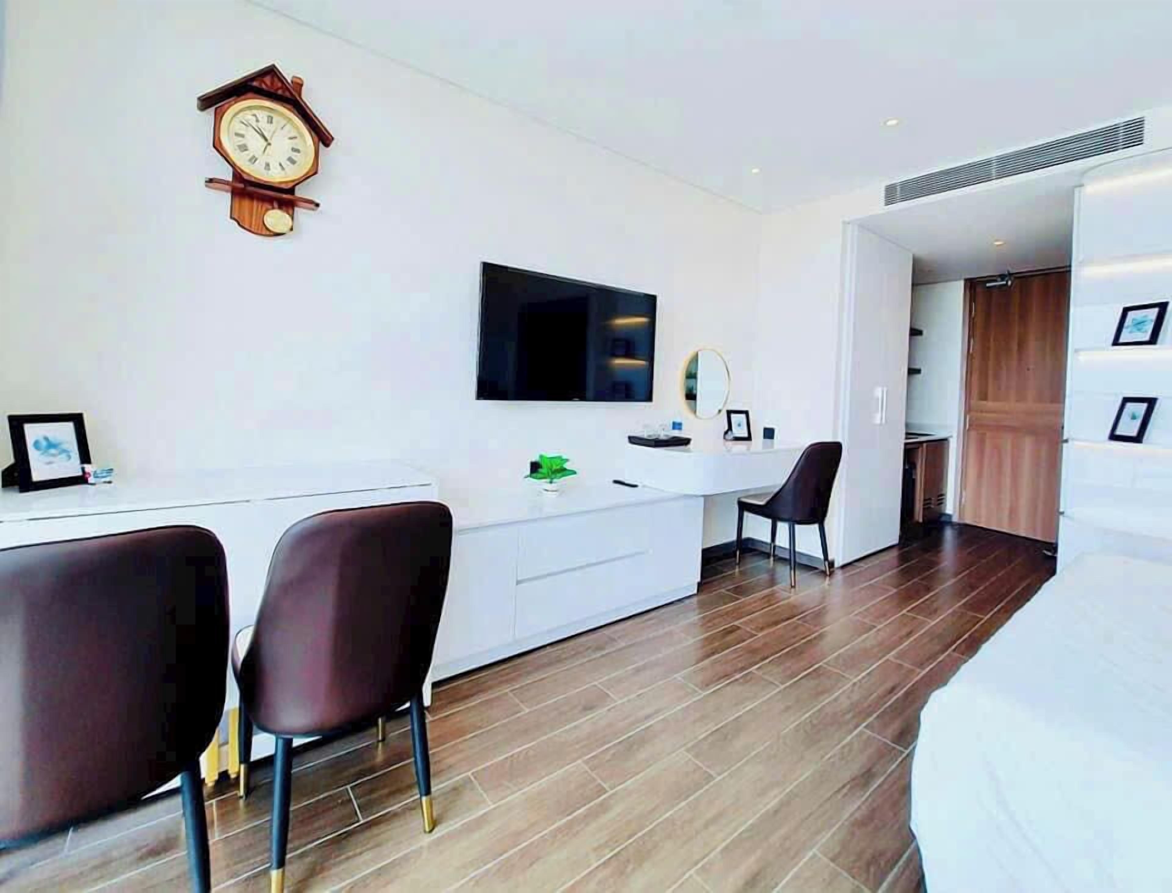 Ein modernes Hotelzimmer mit Holzboden, einem weißen Schreibtisch mit Stuhl, einem großen Flachbildfernseher, einer Uhr und einem Spiegel.