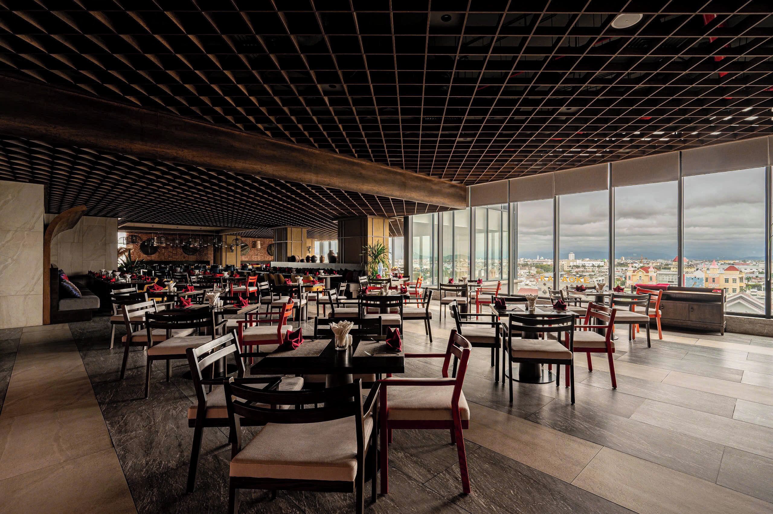 A spacious restaurant with a modern ceiling design, large windows offering a city view, and neatly arranged tables and chairs.