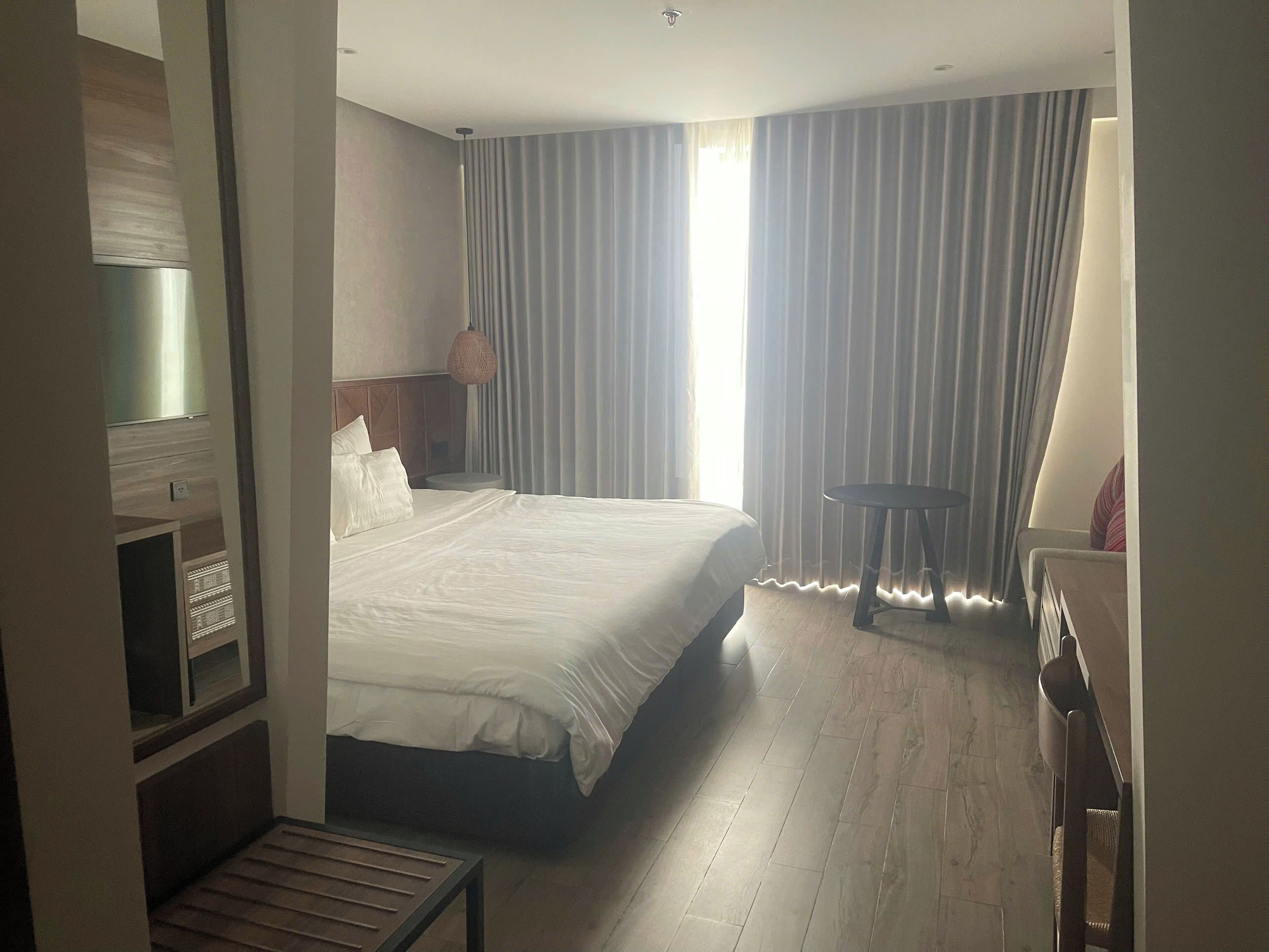 Ein Hotelzimmer mit einem großen Bett, einem kleinen runden Tisch, einem Fernseher und einem Fenster mit Vorhängen.